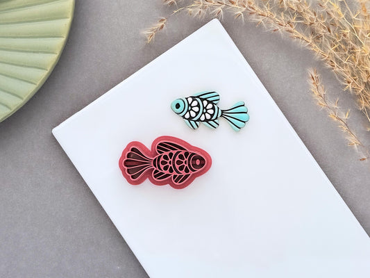 Ornamental fish polymer clay cutter displayed beside a matching pale teal clay cut-out. The folk art-inspired fish design features decorative scales and flowing fins and is photographed on a white ceramic tile.
