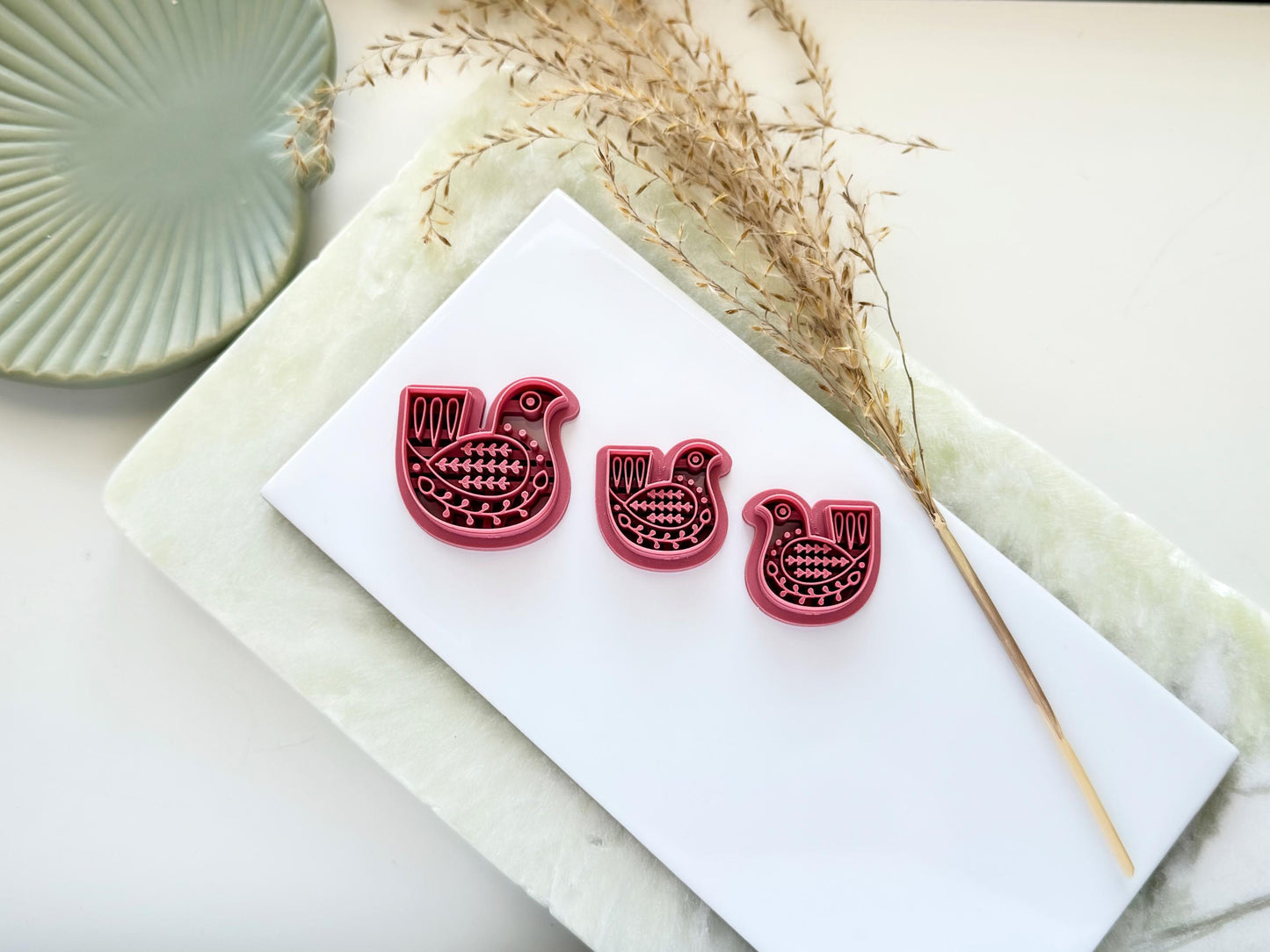 Three pigeon bird shaped clay cutters in pink plastic, two mirrored small pairs and one larger cutter on a white tile background.