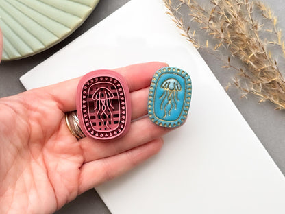 Jellyfish polymer clay cutter shown being held in a hand to demonstrate scale and intricate detail, displayed alongside a finished polymer clay jellyfish sample with an oval beaded frame for creating ocean-inspired jewellery, charms and coastal craft projects.