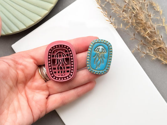 Jellyfish polymer clay cutter shown being held in a hand to demonstrate scale and intricate detail, displayed alongside a finished polymer clay jellyfish sample with an oval beaded frame for creating ocean-inspired jewellery, charms and coastal craft projects.