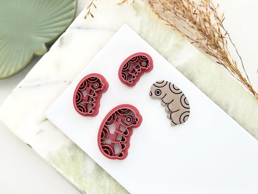 Various sizes of beetle larvae shaped cutter with polymer clay cut out sample showing stamp pattern. All are on a white tile.