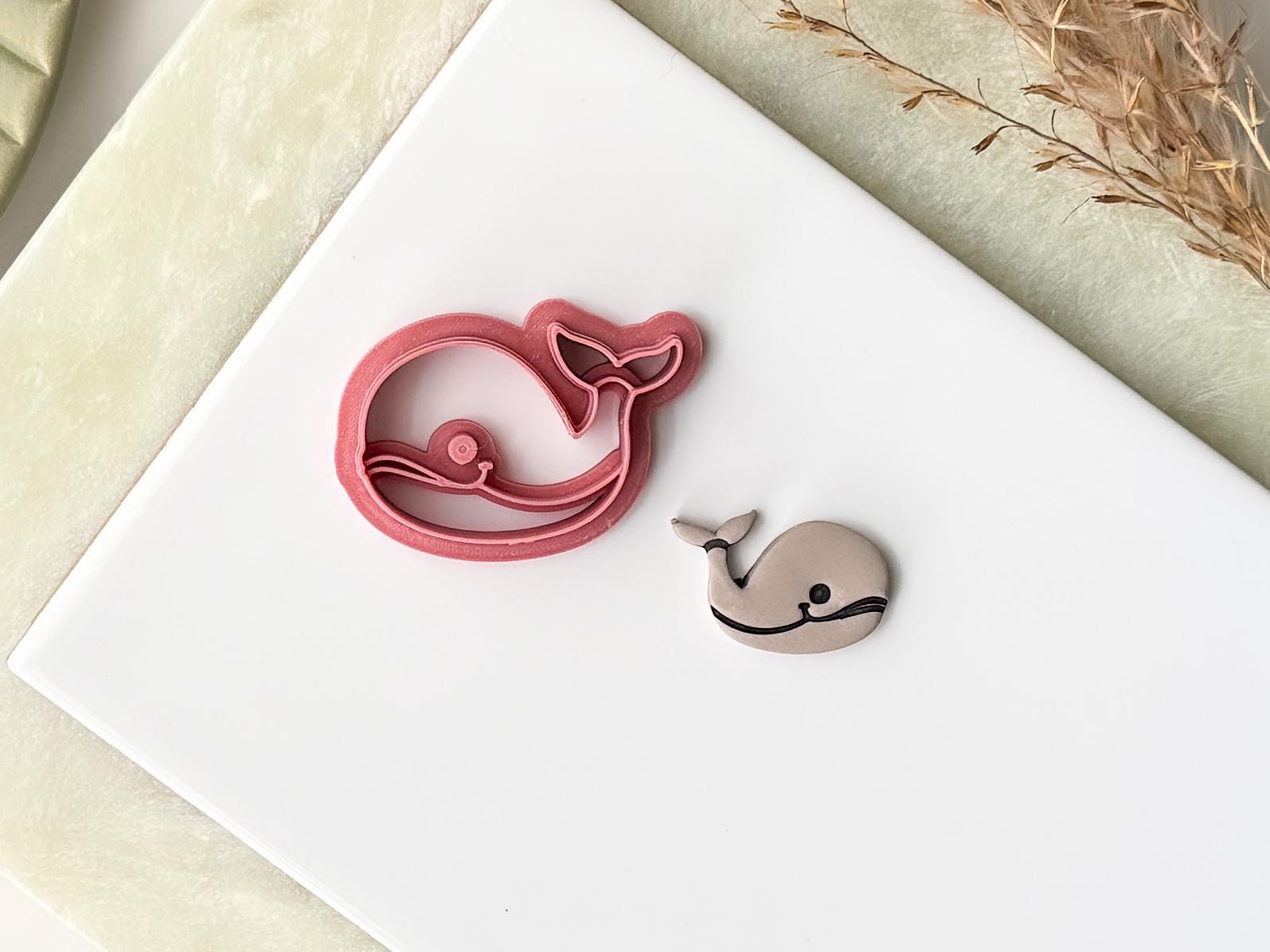 Pink whale shaped craft cutter with smiling face stamp details. Next to the cutter is a whale polymer clay cut out. Both are on a white baking tile.