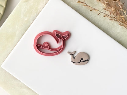 Pink whale shaped craft cutter with smiling face stamp details. Next to the cutter is a whale polymer clay cut out. Both are on a white baking tile.