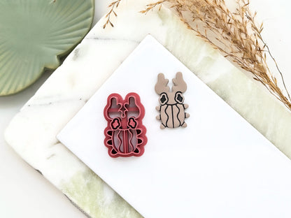 Horn beetle shaped clay cutter with wavy shell, eyes and large horns. Next to the cutter is a polymr clay cut out sample earring