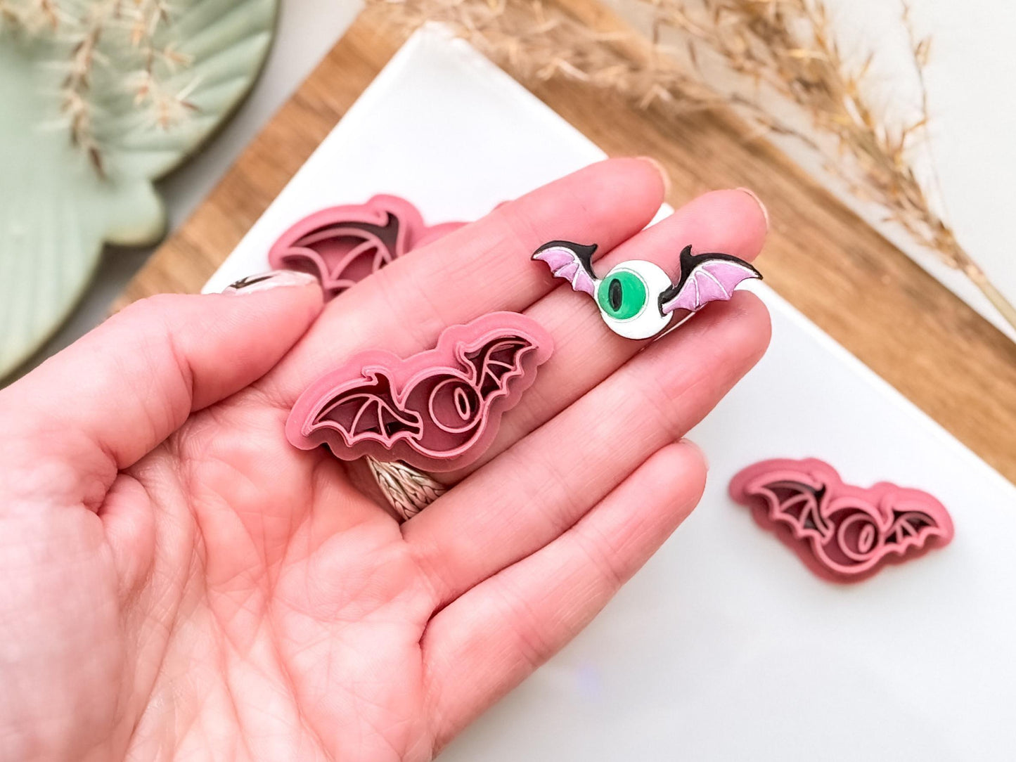 Dark pink flying eye spooky clay cutter in hand next to a painted clay cut out