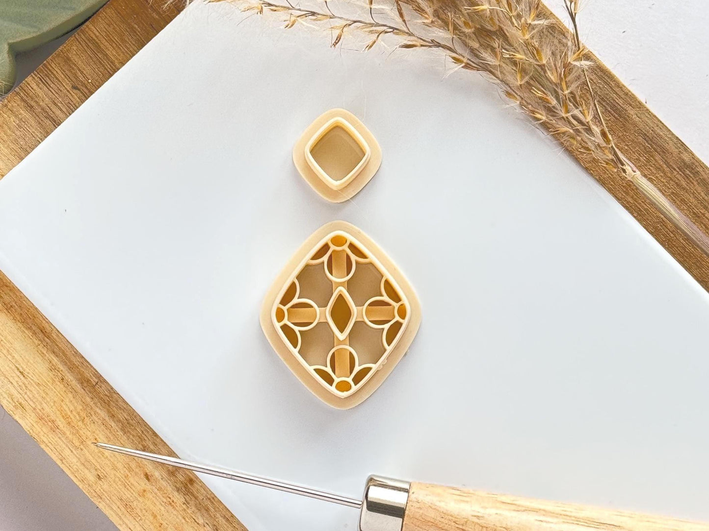 Beige coloured clay earring cutter with flower shaped imprint lines on the main earring cutter piece. Set sits on a narrow white baking tile with dried flowers and a clay tool to the side.
