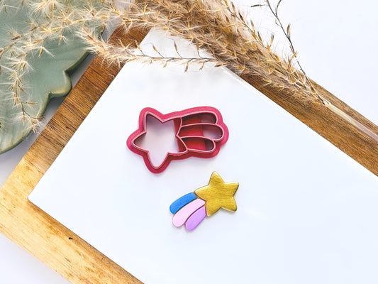 Shooting star shaped clay cutter with five point star and 3 trails separated by imprint lines. Next to the cutter is a gold sample clay cut out with with blue, pink and purple coloured star dust trails