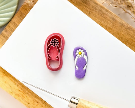 Polymer clay cutter in the shape of a summer flip flop with straps and flower detail next to a purple flipflop cut out clay sample with painted details.