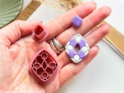 two diamond shaped clay cutters with daisy flower patterns around the outside of the larger dangle piece. Smaller piece is plain and will hold an earring stud pin. Next to this is a purple polymer clay earring created using the clay cutter with the flowers painted white.