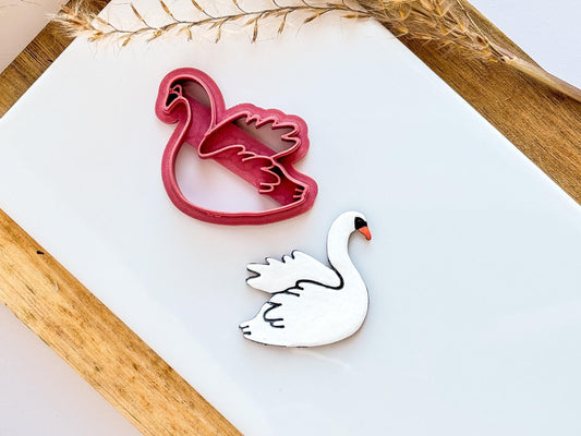 swan clay cutter in dark pink showing detailed stamp and imprint lines, lying next to a polymer clay swan cut out using this swan cutter. Clay sample is painted in white with an orange beak.
