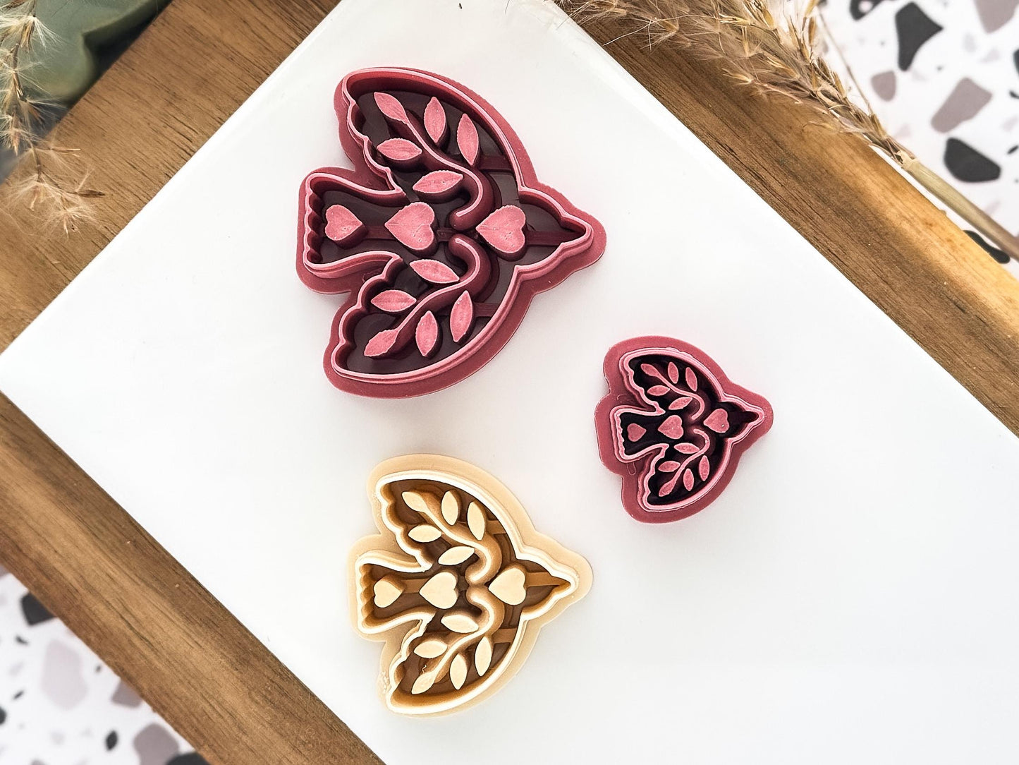 Three bird-shaped clay cutters in varying sizes. The cutters are in shades of pink and beige, with intricate leaf and heart designs. The cutters are arranged on a white surface.