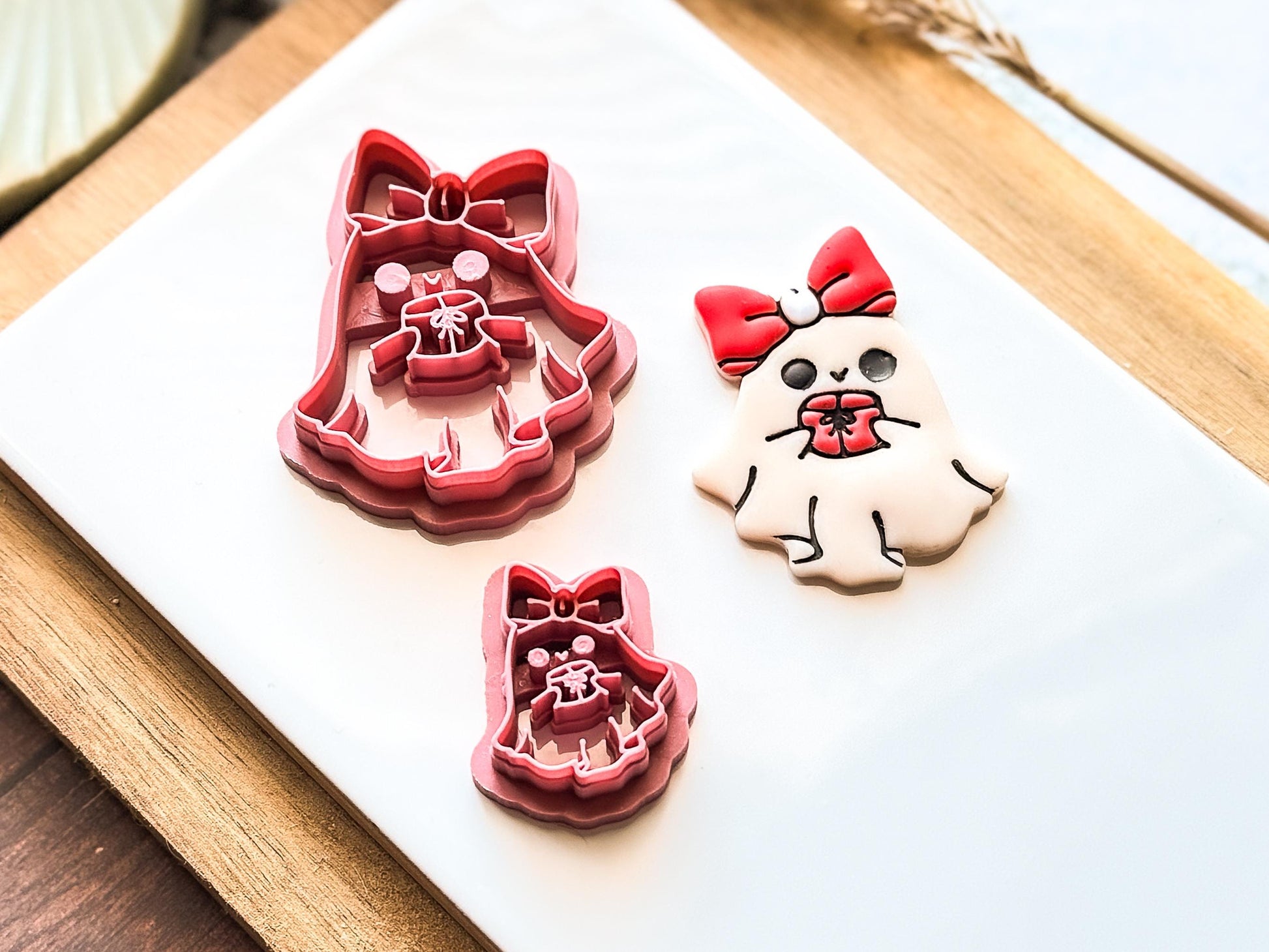 Three clay cutters and a decorated clay earring in the shape of a ghost with a red bow and a gift. The cutters are pink and the polymer clay cut-out is white with black details.