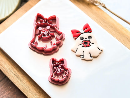 Three clay cutters and a decorated clay earring in the shape of a ghost with a red bow and a gift. The cutters are pink and the polymer clay cut-out is white with black details.