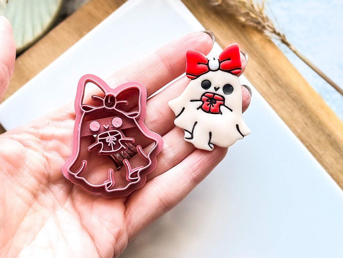 A pink ghost-shaped clay cutter and a white ghost keyring with a red bow and gift. The clay cutter is a ghost with a bow, holding a gift. The keyring ghost has a red bow and is holding a red gift.
