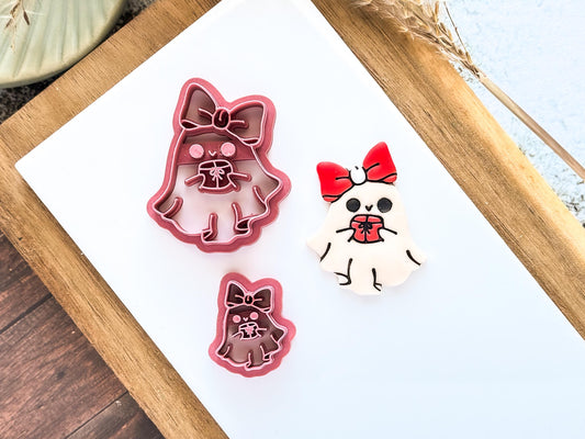 Ghost-shaped clay cutters and a painted polymer clay cut out. The clay cutters are pink and feature a bow on top. The cut-out is white with a red bow and a gift.