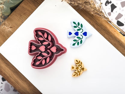 A bird-shaped clay cutters on a white surface. One is a dark red cutter with leaf and heart details. Along side the cutters are two polymer clay cut out samples.