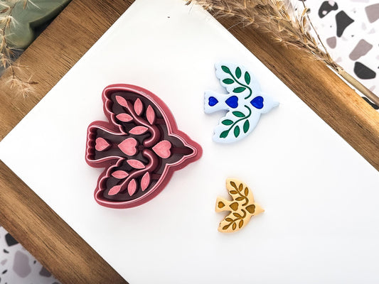 A bird-shaped clay cutters on a white surface. One is a dark red cutter with leaf and heart details. Along side the cutters are two polymer clay cut out samples.