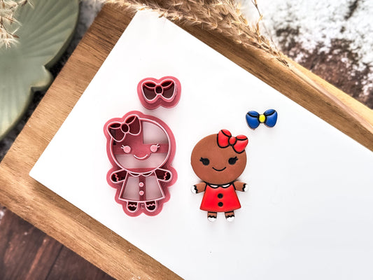 Pink gingerbread girl clay cutter with separate bow cutter on a white tile with a polymer clay sample cut out. Sample has been painted with liquid clay in Christmas themed colours.