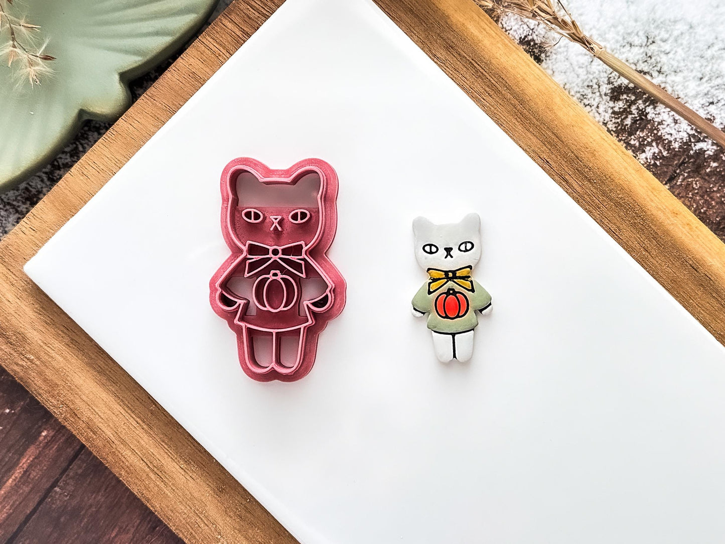 pink cat shaped polymer clay cutter with bow and pumpkin jumper details on a baking tile with a white and green clay cut-out sample.