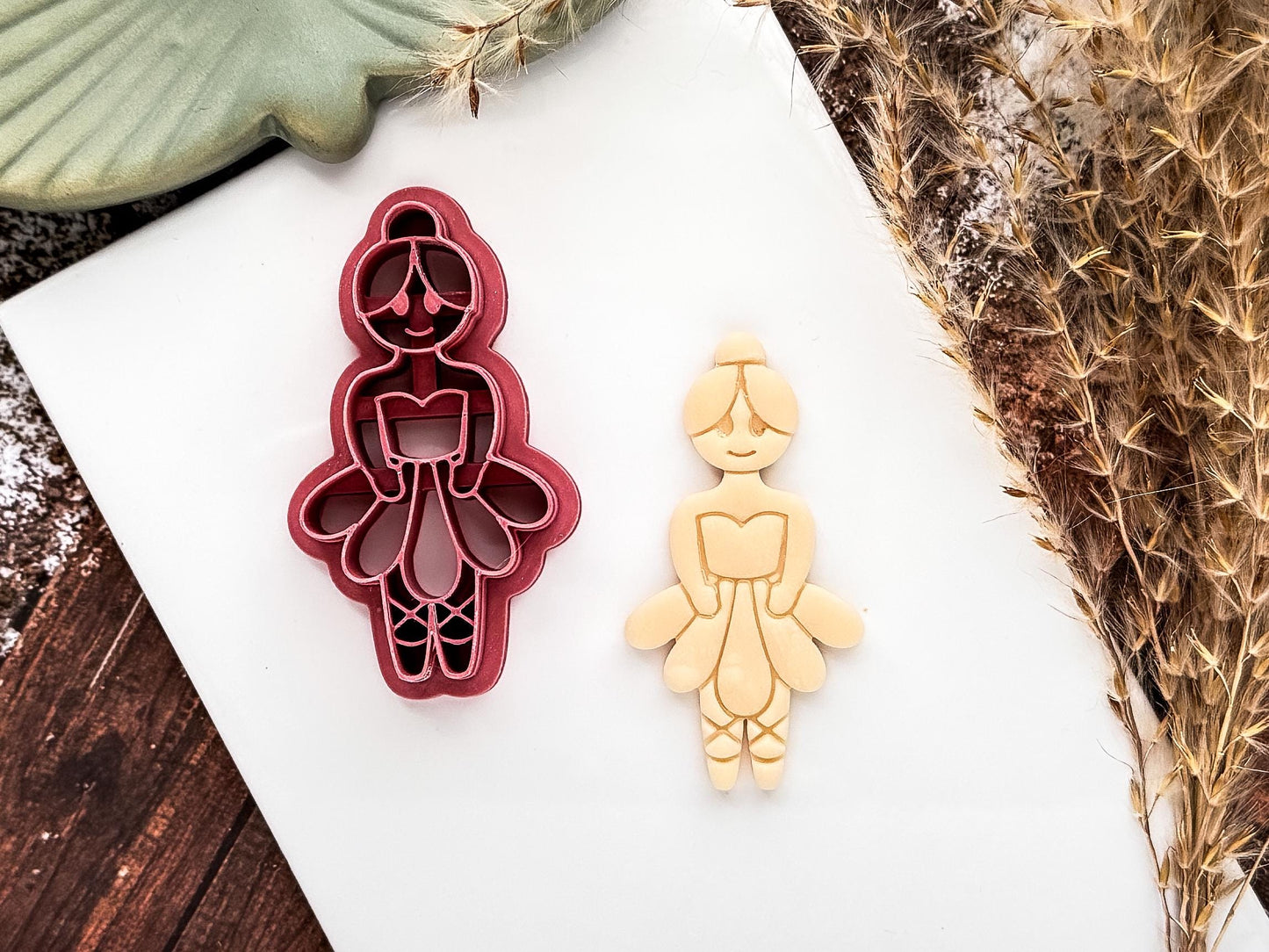 Pink ballerina shaped polymer clay cutter with tutu, ballet slippers and bun details. Beige cut out sample of polymer clay is next to the cutter on a white tile.