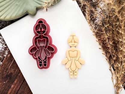 Pink ballerina shaped polymer clay cutter with tutu, ballet slippers and bun details. Beige cut out sample of polymer clay is next to the cutter on a white tile.