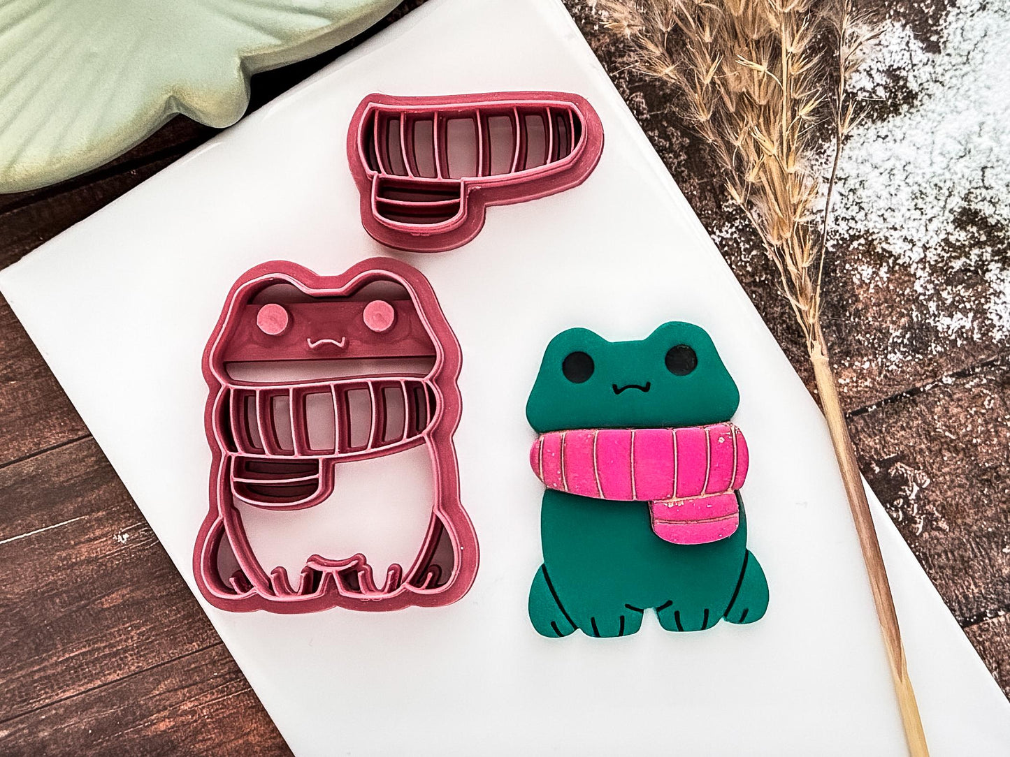 Frog shaped polymer clay cutter with separate scarf along with green polymer clay sample cut out on a white clay tile