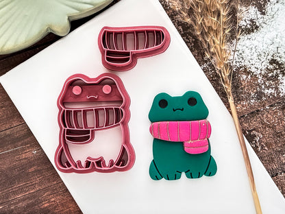 Frog shaped polymer clay cutter with separate scarf along with green polymer clay sample cut out on a white clay tile