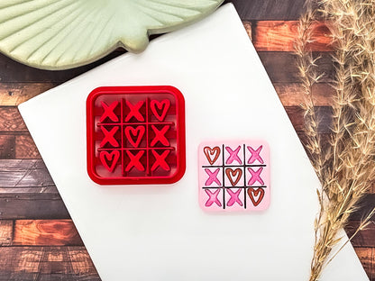 Red hearts and crosses game shaped clay cutter with debossing hearts and crosses details lying next to a pink polymer clay sample painted with metallic pink and copper acrylic paint.