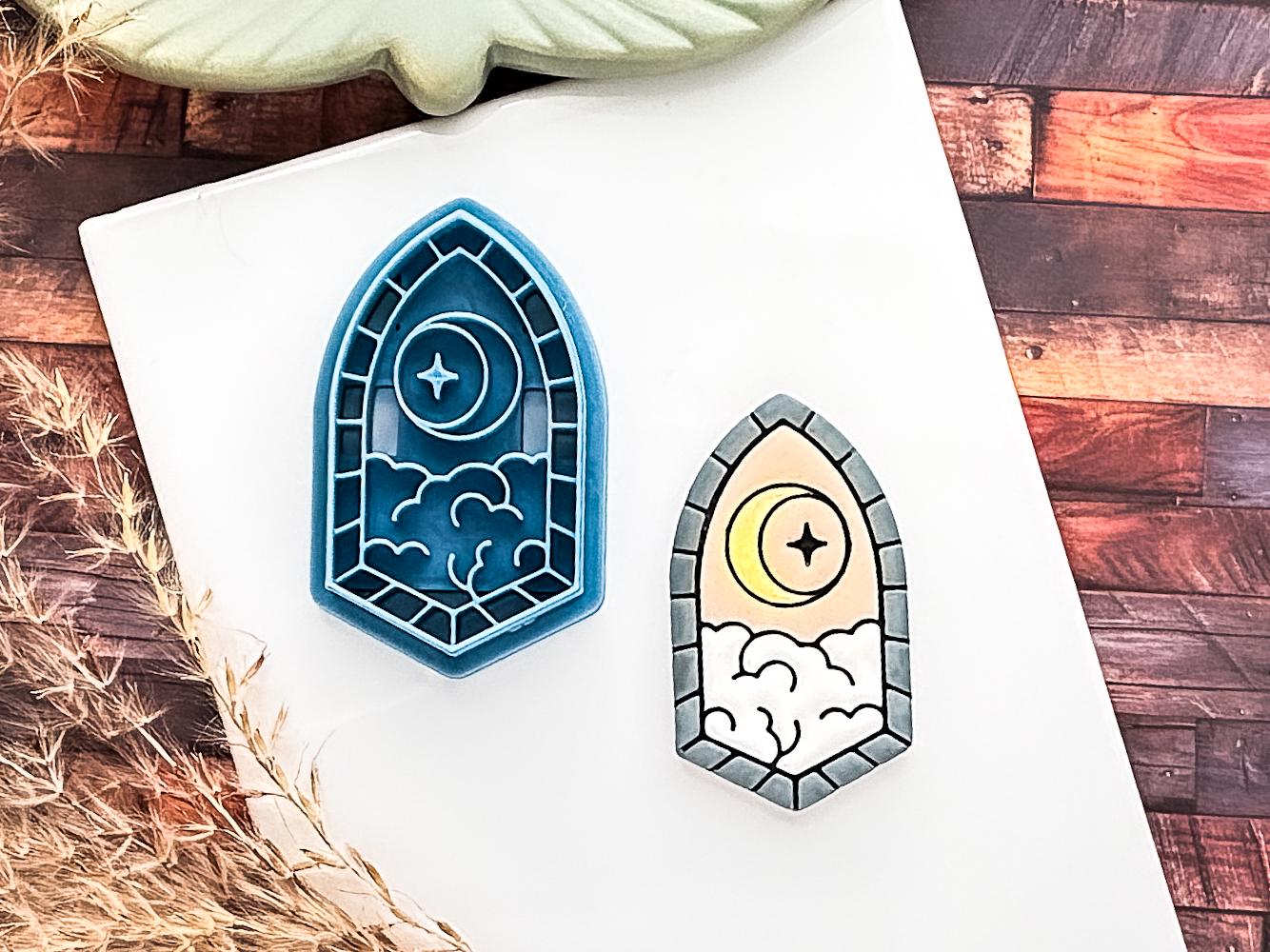 blue clay cutter in the shape of a pointed window with brick details on the window frame and a moon, star and clouds internal stamp design. Painted polymer clay cut out sample is beside it sitting on a white baking tile.
