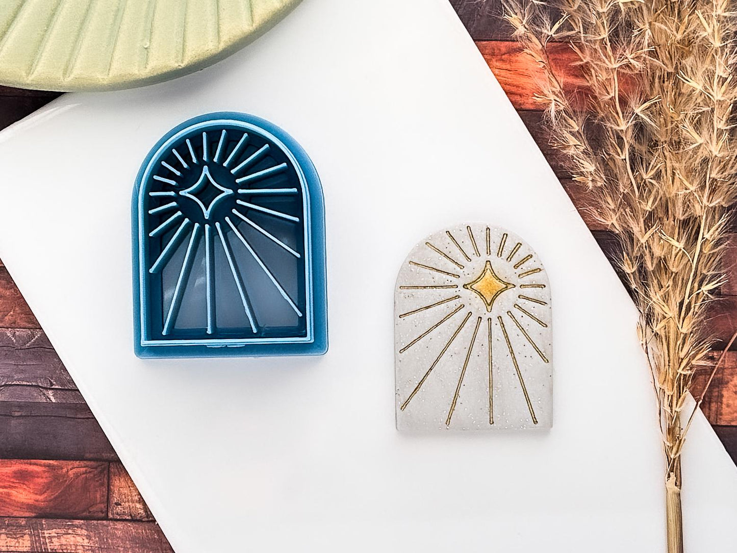 Celestial themed arch shaped clay cutter with radiating beam lines and off-centre 4 point star in blue PLA material. Next to it is a sampe polymer clay cut out using glitter silver fimo clay and decorated using gold acrylic paint.