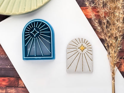 Celestial themed arch shaped clay cutter with radiating beam lines and off-centre 4 point star in blue PLA material. Next to it is a sampe polymer clay cut out using glitter silver fimo clay and decorated using gold acrylic paint.