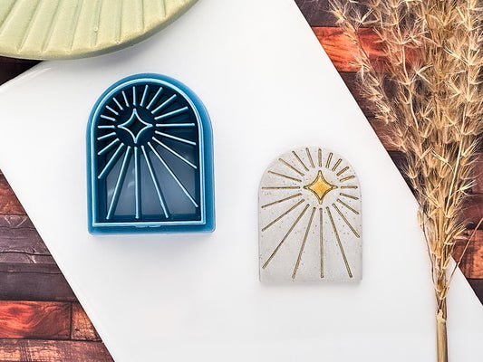 Celestial themed arch shaped clay cutter with radiating beam lines and off-centre 4 point star in blue PLA material. Next to it is a sampe polymer clay cut out using glitter silver fimo clay and decorated using gold acrylic paint.
