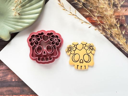 Skull shaped clay cutter with flower and heart details next to a beige polymer clay cut out painted with acrylic paint to show the stamp details.
