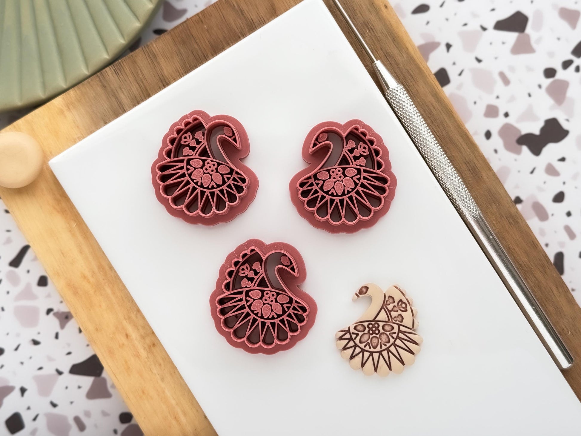 3 scandi style swan clay cutters with folk floral stamp patterns. It includes a mirrored pair for clay earring making and single cutter for pendant making and fondant decoration making. Next to these are a beige clay sample cut out.