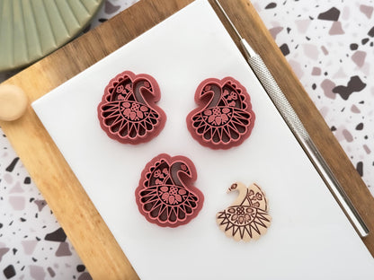 3 scandi style swan clay cutters with folk floral stamp patterns. It includes a mirrored pair for clay earring making and single cutter for pendant making and fondant decoration making. Next to these are a beige clay sample cut out.