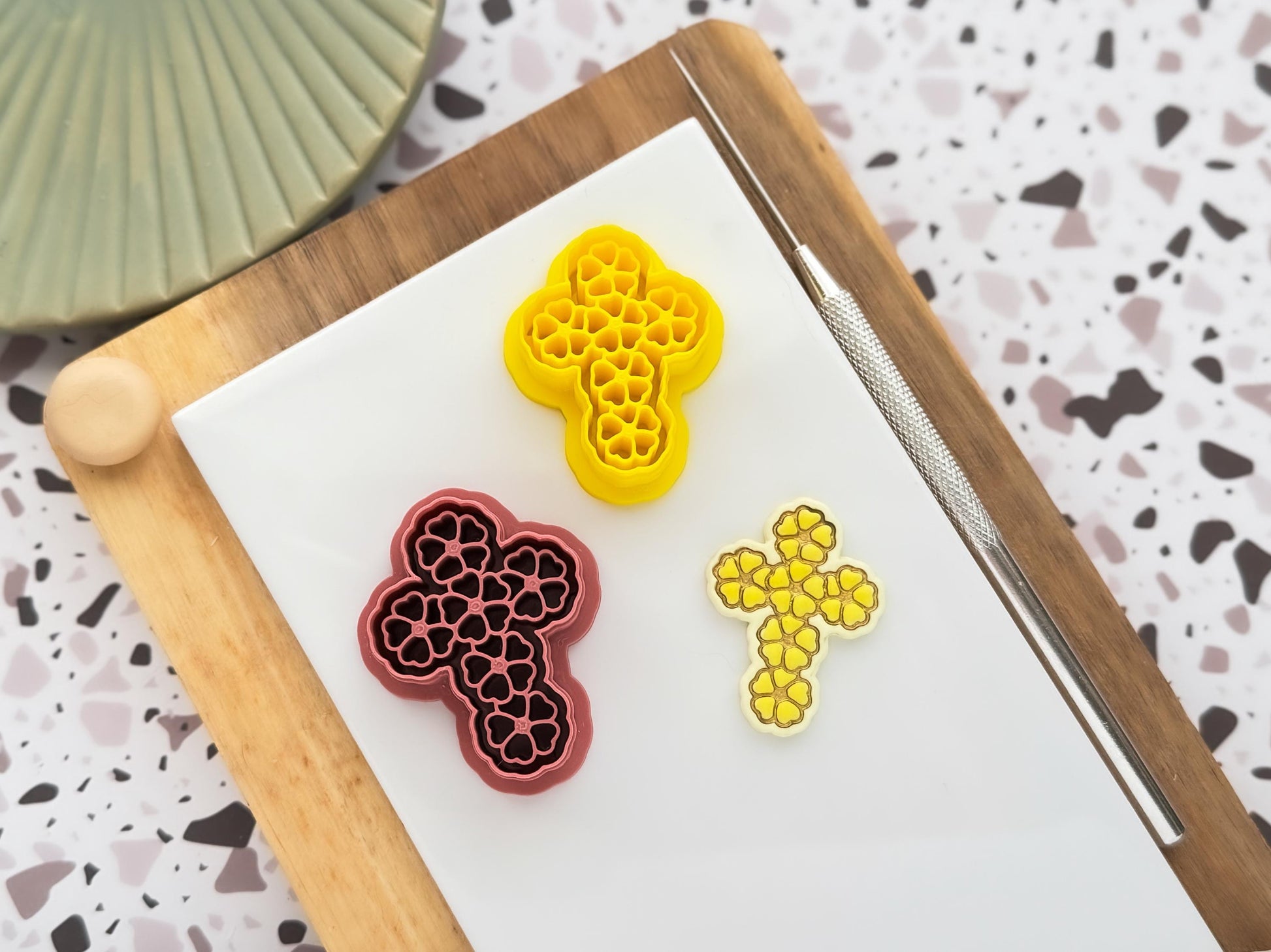 Images shows two cross shaped clay cutters with daisy flower stamp details in pink and yellow, beside them is a pastel yellow polymer clay cut out sample piece on a white baking tile and clay tools.