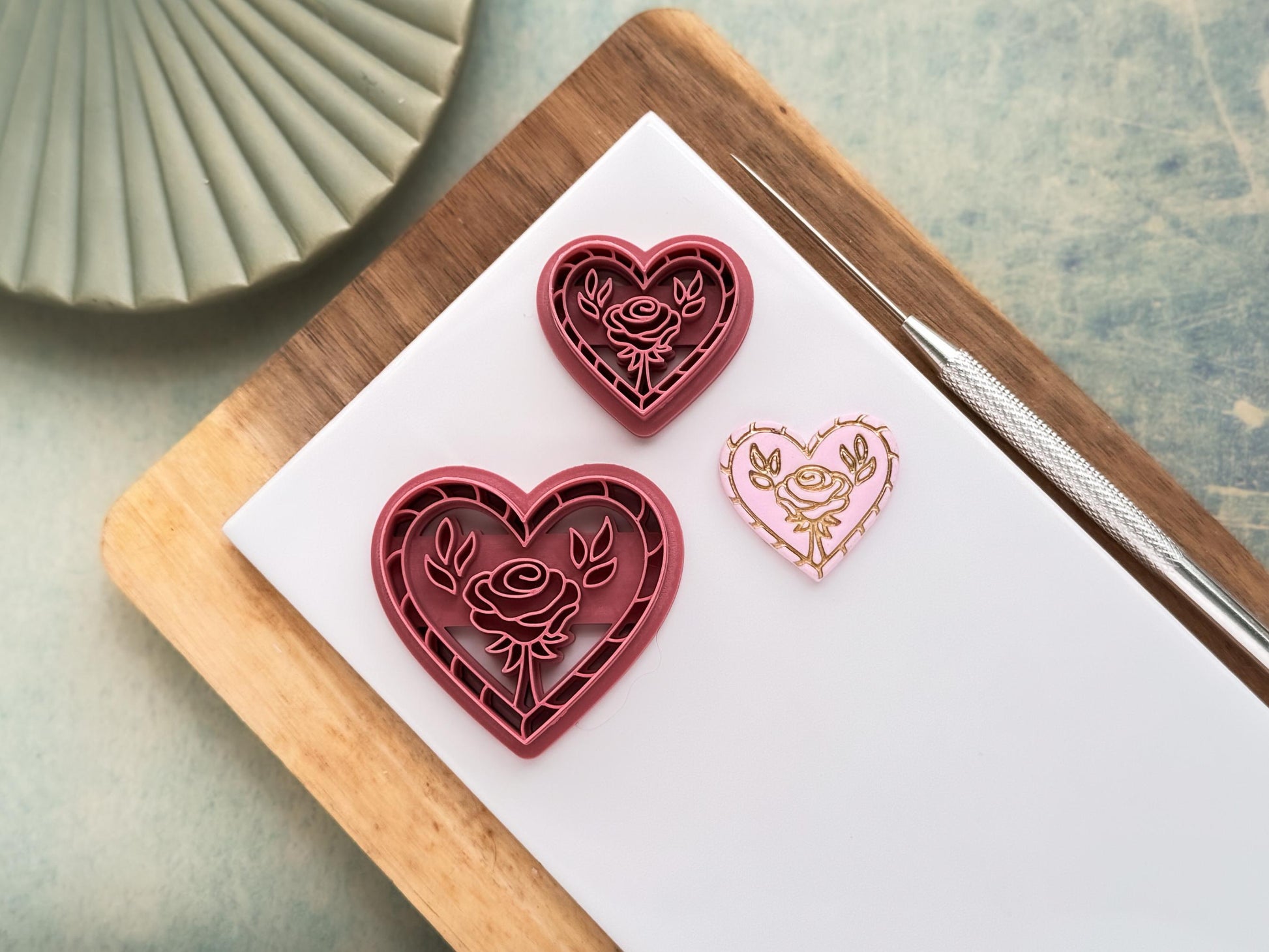 this image contains two pink polymer clay cutters in a heart shape with rope pattern outer edge detail and a rose flower with leaves in the middle. One cutter is 3cm and the other is 4 cm. Next to the cutters is a pastel pink polymer clay sample cut out with the stamped flower details painted in gold.