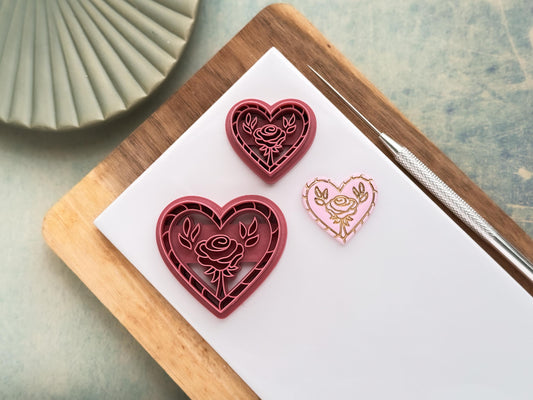 this image contains two pink polymer clay cutters in a heart shape with rope pattern outer edge detail and a rose flower with leaves in the middle. One cutter is 3cm and the other is 4 cm. Next to the cutters is a pastel pink polymer clay sample cut out with the stamped flower details painted in gold.