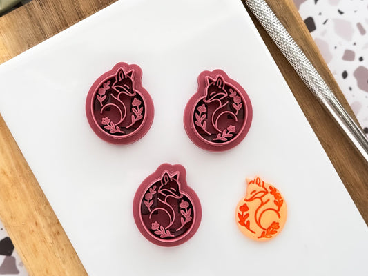 Scandi style folk curled fox shaped clay cutter in mirrored pair and single cutter. Orange faux ceramic decorated polymer clay cut out shape lies next to them.