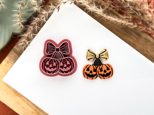 pink halloween clay cutter in the shape of a pair of pumpkin cherries. Each pumpkin features debossed triangle eyes and nose and jagged smile, the cherry stems and a ribbon tied with a bow.