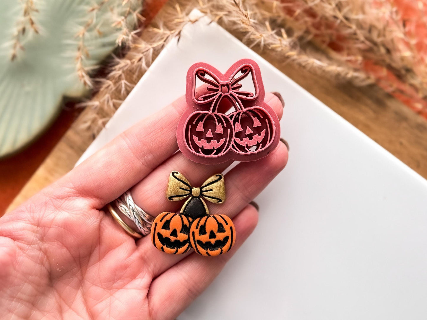 cherry pumpkin clay cutter with scary face and bow details next to a polymer clay cut out painted in orange and gold. Both a being held in a hand for scale.