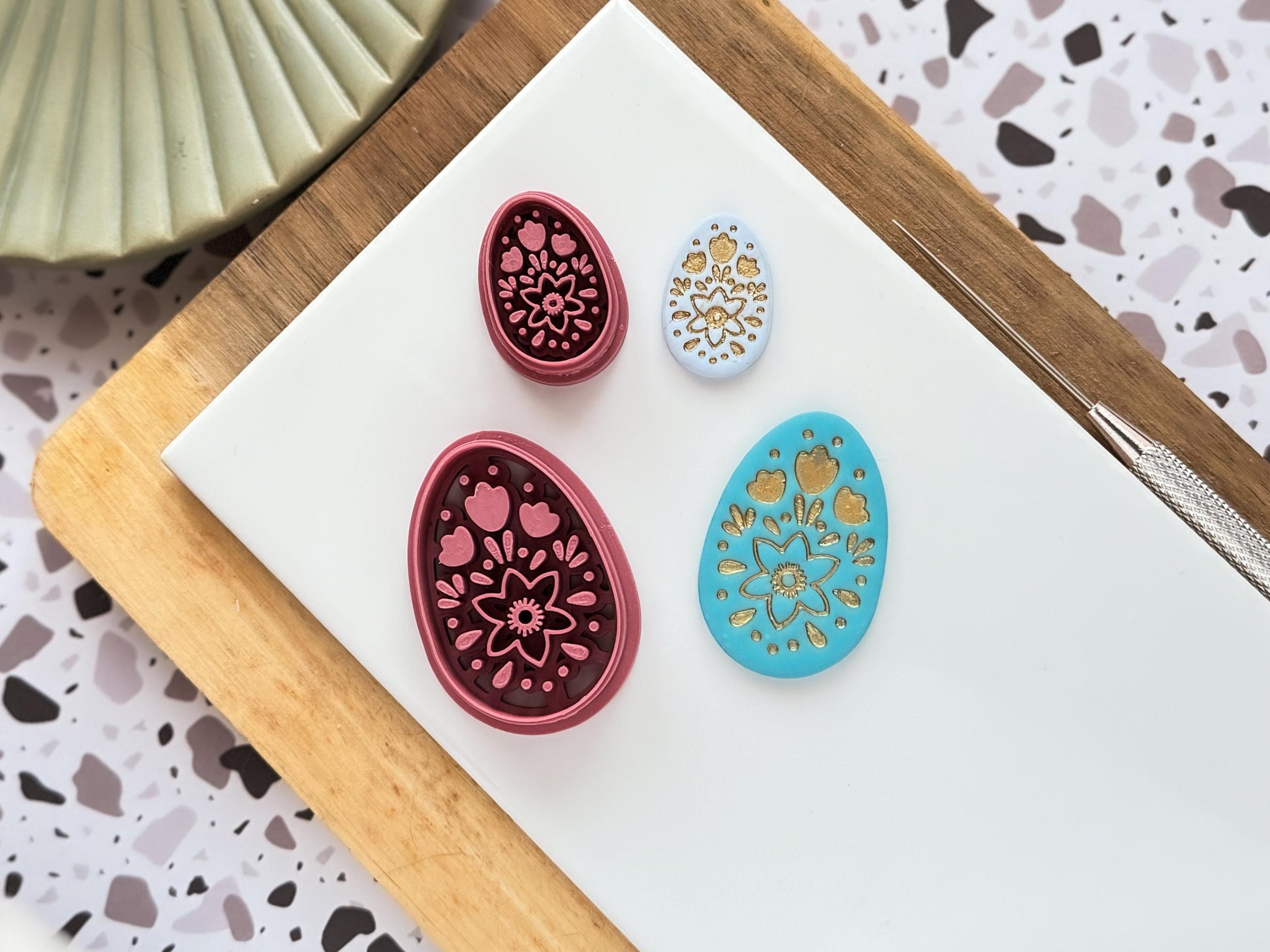 Boho folk art style easter egg shaped clay cutter in large and small sizes. Next to them are polymer clay samples with gold details on a baking tile.