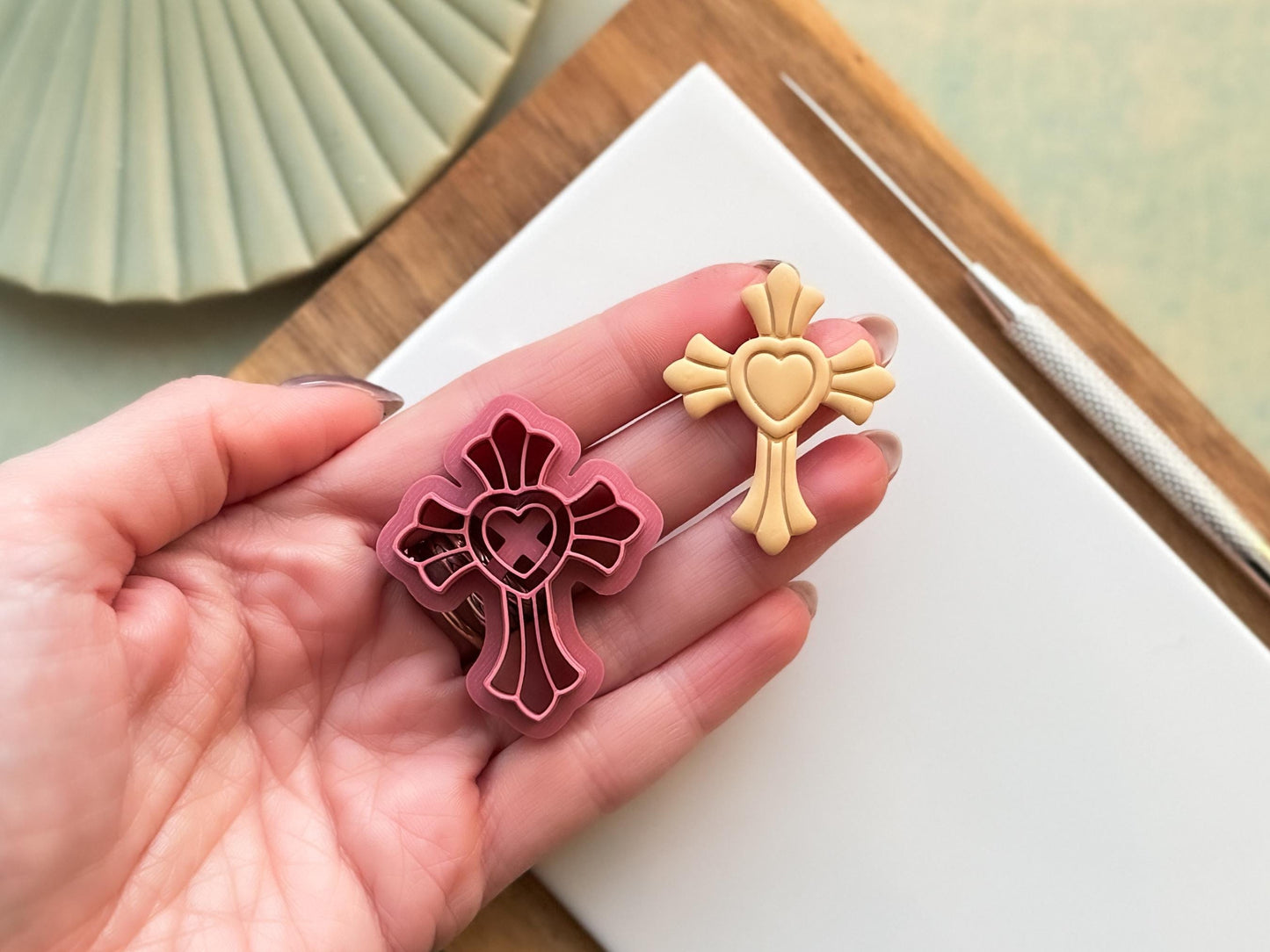 Cross clay cutter with scalloped edges and heart shaped middle, next to it is a beige polymer clay cut out sample being held in a hand for scale.