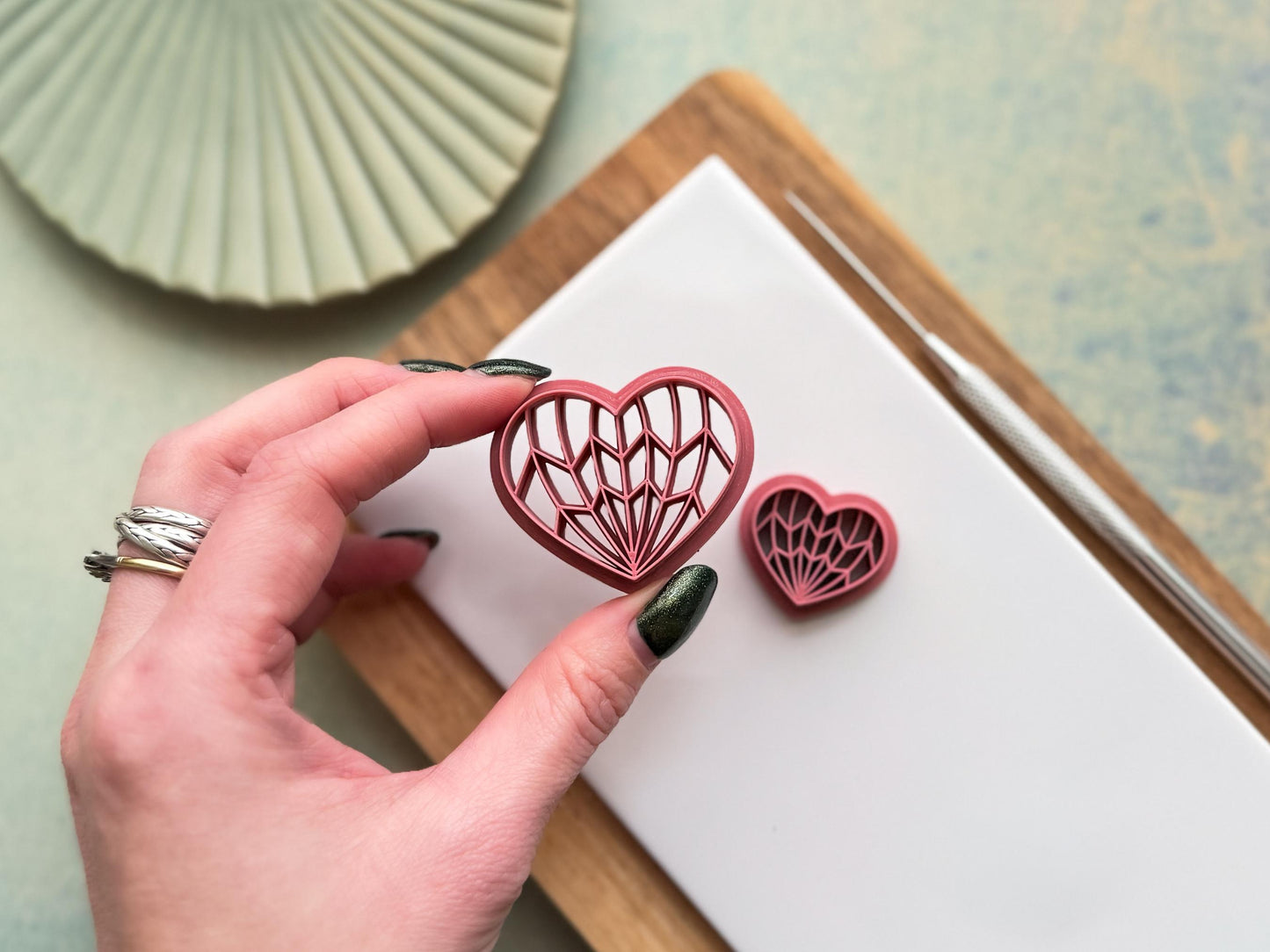Hand holding geometric heart polymer clay cutter with intricate Art Deco line pattern, showing size and detail on clay workspace