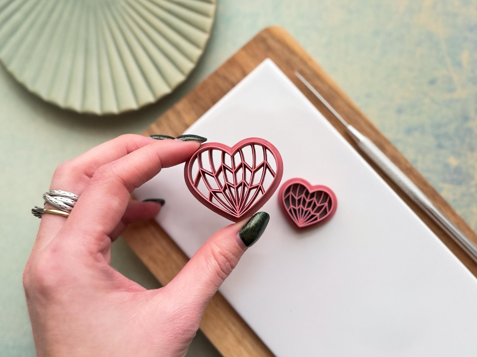 Hand holding geometric heart polymer clay cutter with intricate Art Deco line pattern, showing size and detail on clay workspace