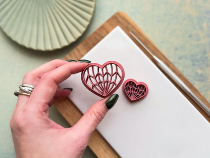 Hand holding geometric heart polymer clay cutter with intricate Art Deco line pattern, showing size and detail on clay workspace