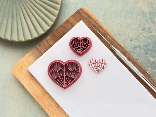 Two geometric heart polymer clay cutters with Art Deco style line pattern, shown with a finished pink and gold clay piece on a white tile workspace.