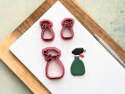 Small and large spray bottle shaped clay cutter in pink. One cutter is a single shape, the other is a mirrored pair for clay earring making. Next to these are a polymer clay cut out sample. All are resting on a white clay baking tile.