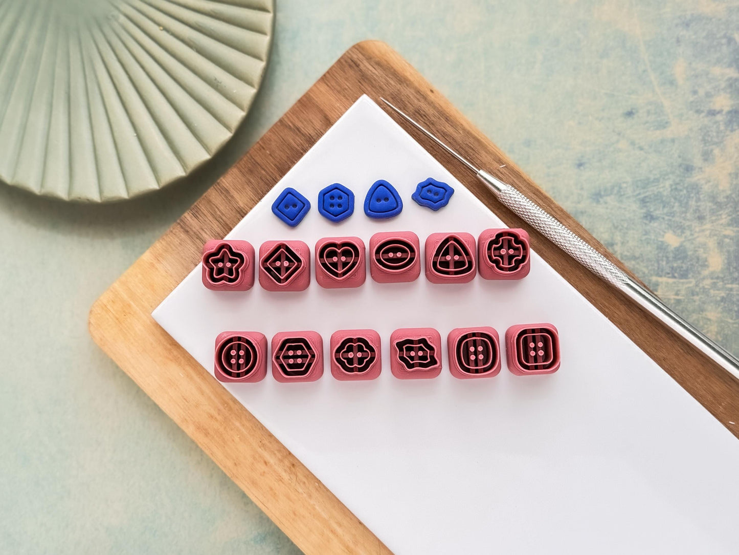 Set of 12 small clay cutters in the shape of buttons with button hole details. Next to the cutter set is four blue polymer clay sample pieces.
