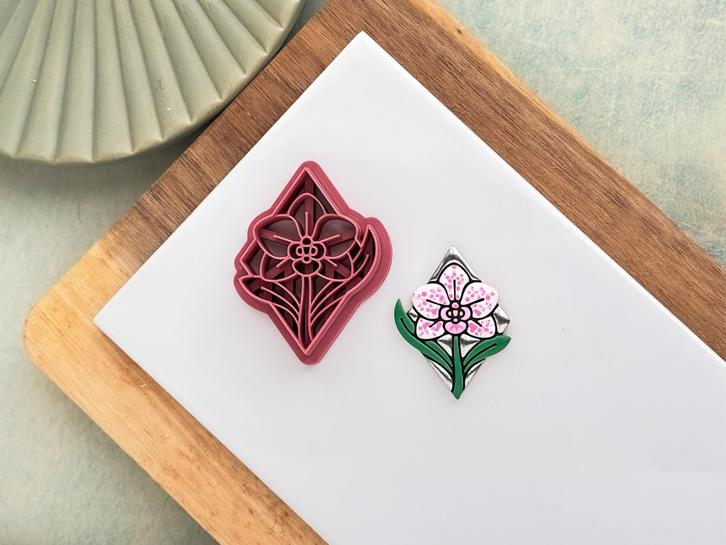 Pink clay cutter in the shape of a diamond with orchid flower and leaf stamp details. Next to it is a polymer clay sample cut out painted in chrome silver, green, white, and pink.